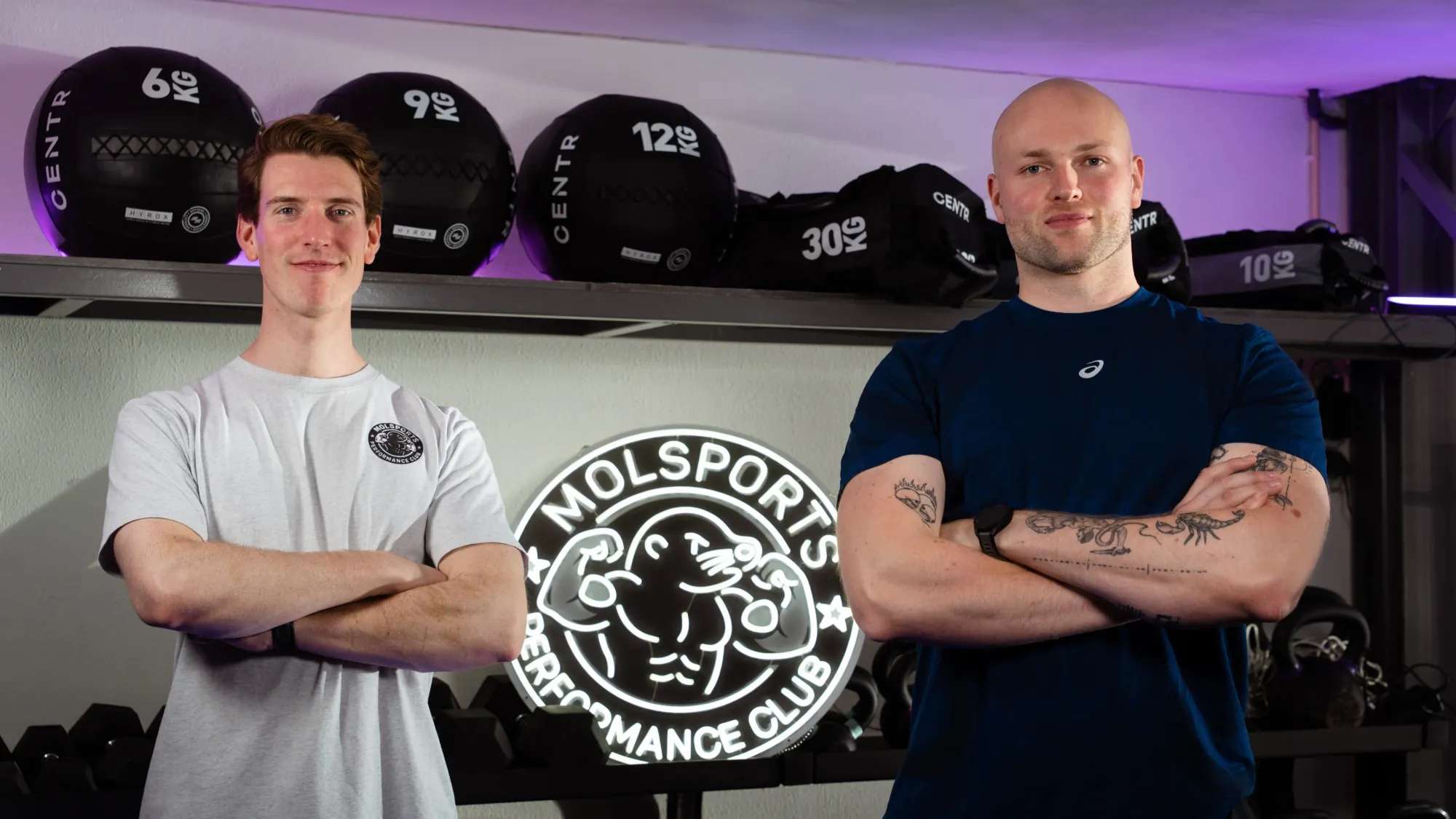 Coaches Nick en Danny voor het lichtgevende MOLSPORTS-logo in de zaal